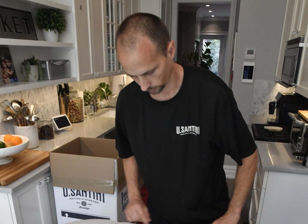A man wearing a black shirt is unpacking a box in a modern kitchen. 
