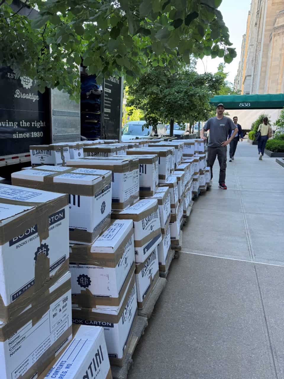Dozens of U. Santini Moving & Storage branded cardboard boxes stacked in long rows on a Manhattan sidewalk beside a moving truck, with a crew member standing among them