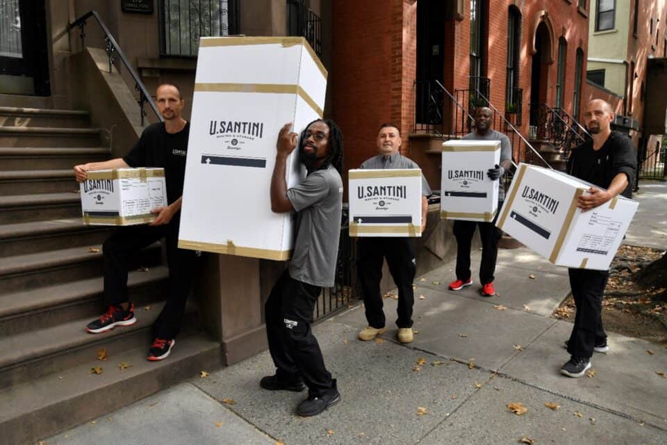 Five U. Santini Moving & Storage crew members holding large branded boxes on the sidewalk outside a row of Brooklyn brownstone buildings