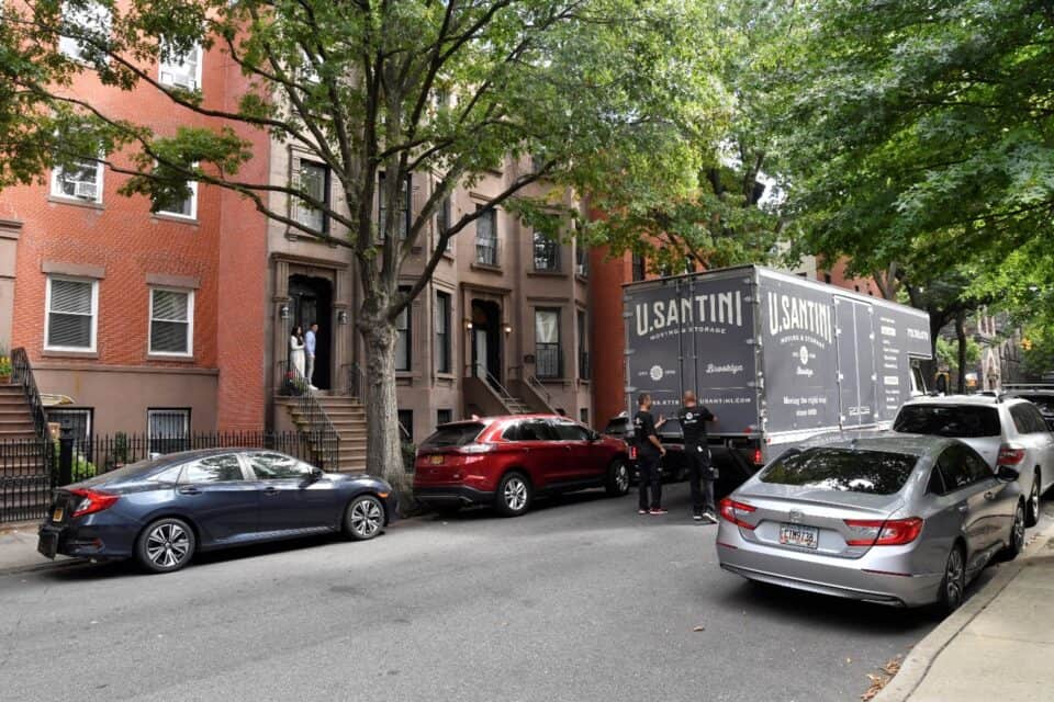 A U. Santini Moving & Storage truck parked on a tree-lined Brooklyn street in front of brownstone buildings, with two customers watching from a front stoop and crew members near the truck