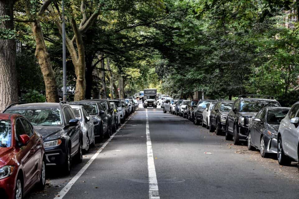 A U. Santini Moving & Storage truck driving toward the camera at the far end of a wide, tree-lined New York City street with cars parked on both sides