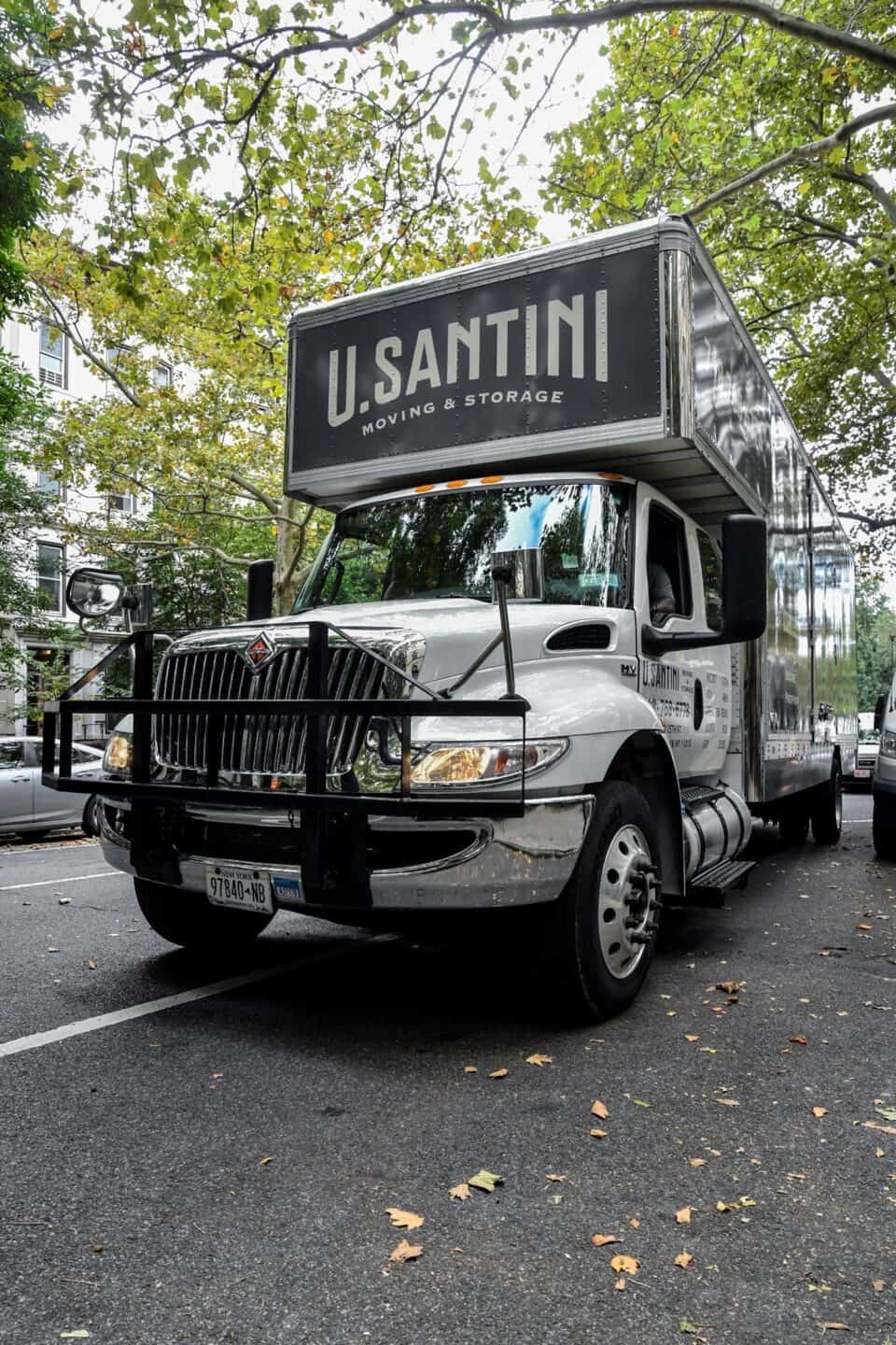 Front view of a U. Santini Moving & Storage truck parked on a street beneath a lush green tree canopy in autumn, with the branded dark gray box clearly visible