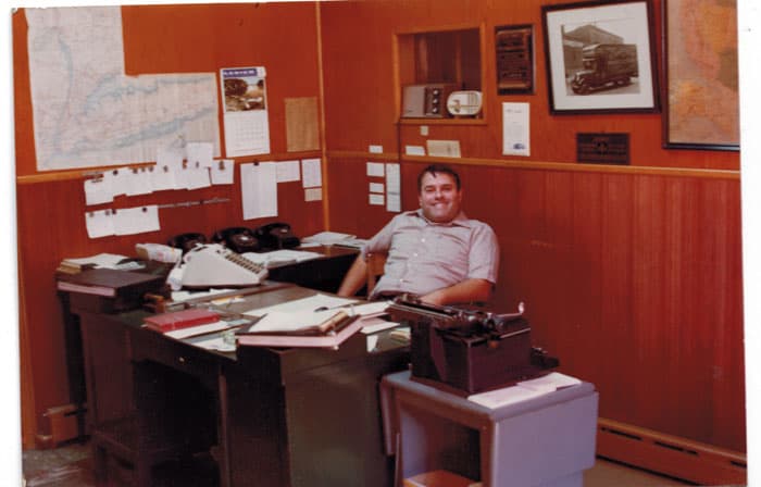 A vintage color photograph of a U. Santini employee smiling at a desk surrounded by papers, phones, and a typewriter in a wood-paneled office, with a framed photo of a company truck on the wall