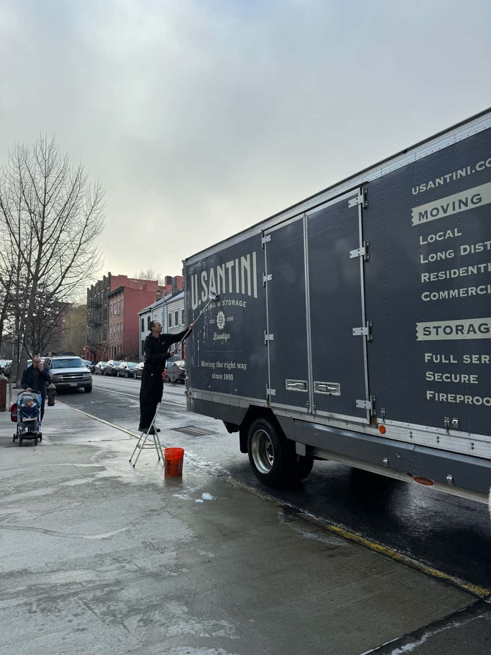 A U. Santini Moving & Storage employee standing on a stepladder to wash the side of a large branded moving truck parked on a wet Brooklyn street on a gray day