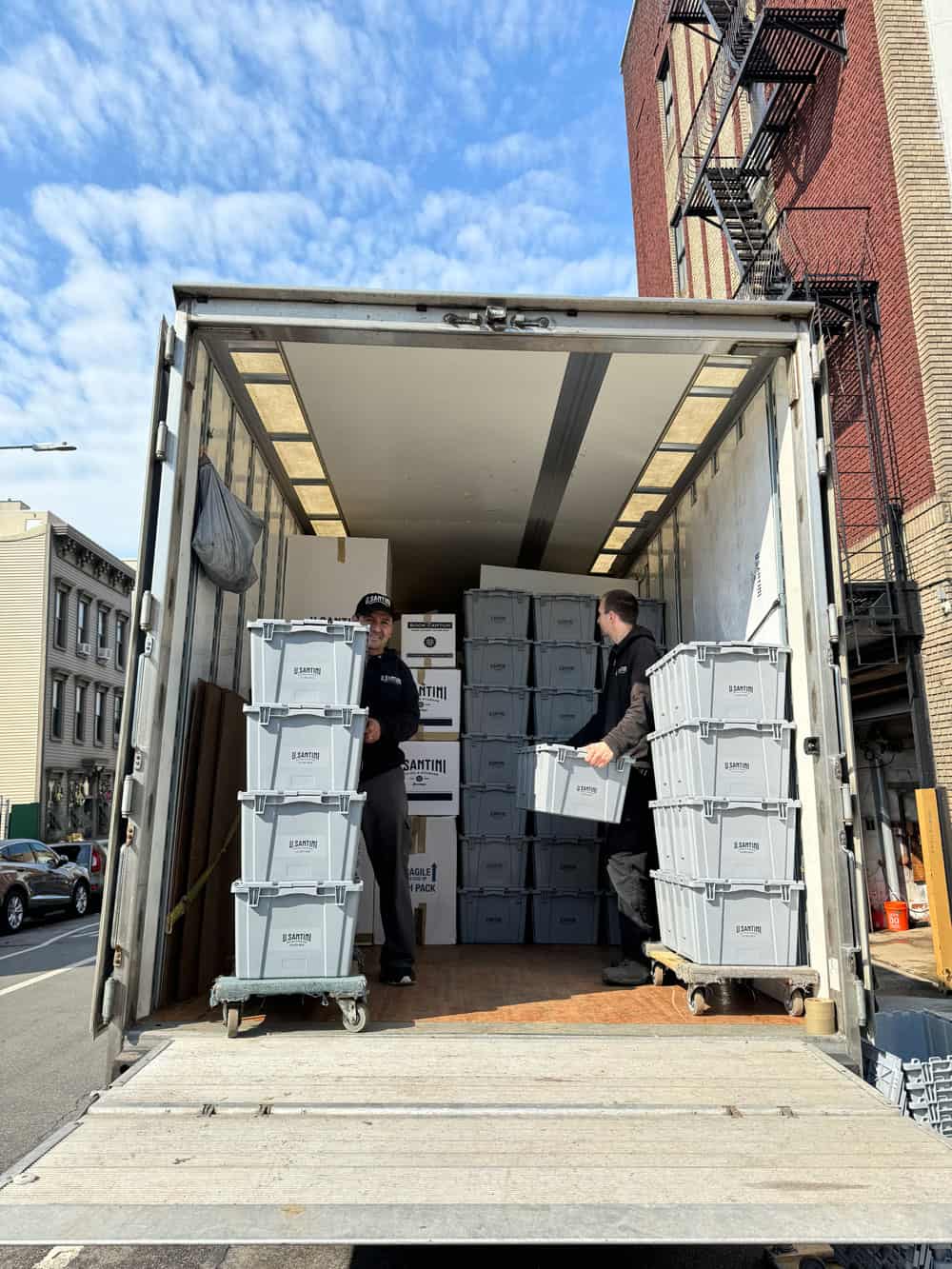 Two U. Santini Moving & Storage crew members unloading tall stacks of branded gray plastic moving bins from the open rear of a truck on a sunny Brooklyn street
