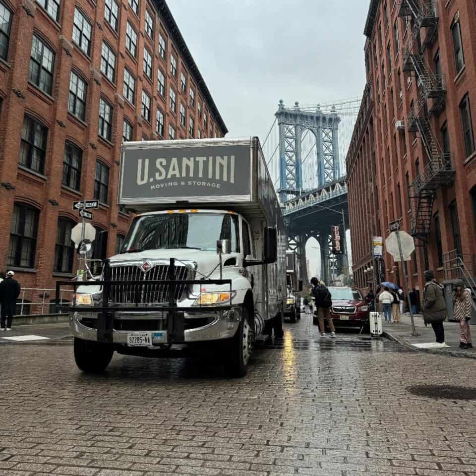 A U. Santini Moving & Storage truck on wet cobblestone streets in the DUMBO neighborhood of Brooklyn, with the iconic Manhattan Bridge framed between brick warehouse buildings in the background