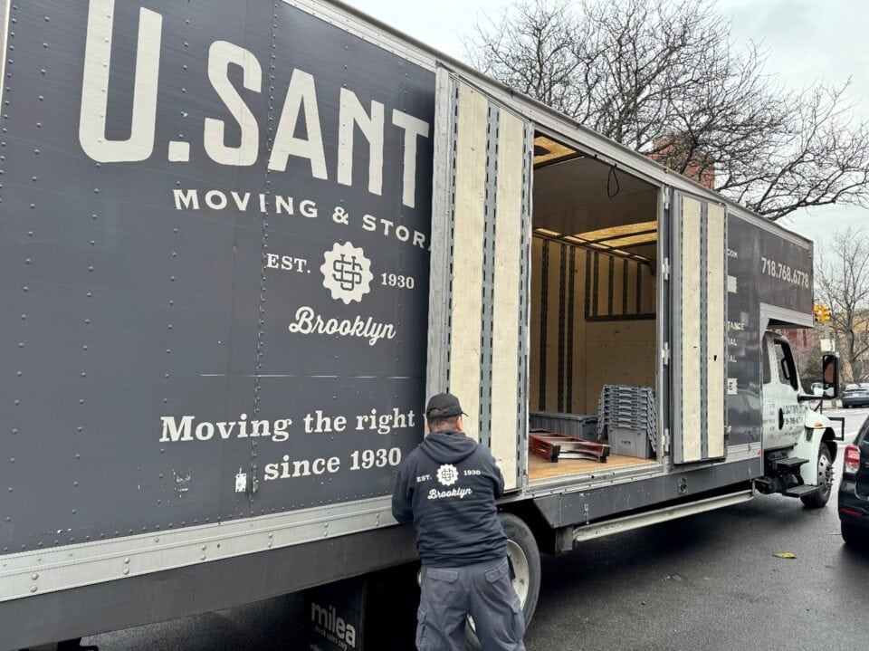 A convoy of U. Santini Moving & Storage trucks driving down a sunny Brooklyn street with brownstone buildings and parked cars on both sides