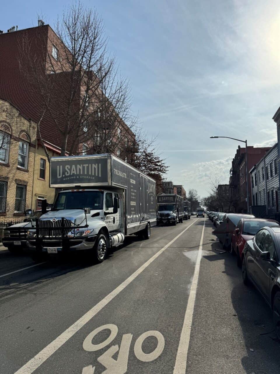 Two U. Santini Moving & Storage trucks driving in convoy through a New York City street lined with brick apartment buildings and parked cars