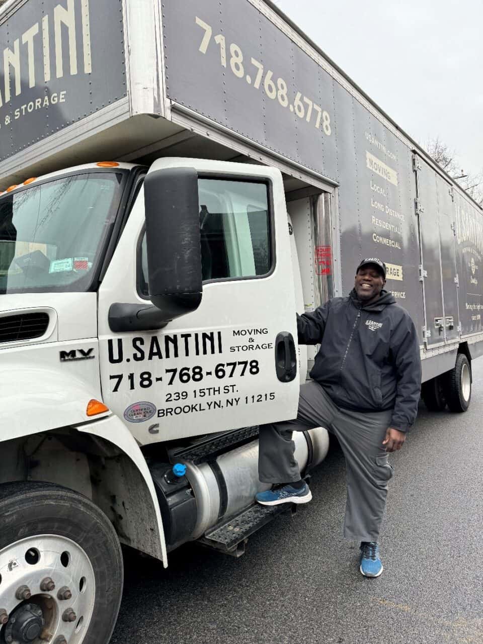 A smiling U. Santini Moving & Storage employee standing beside a company truck displaying the address 239 15th St., Brooklyn, NY 11215 and phone number 718-768-6778