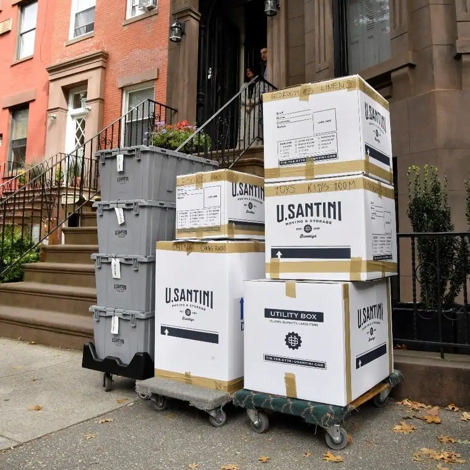 Stacked U. Santini Moving & Storage branded cardboard boxes and gray plastic bins on dollies in front of a Brooklyn brownstone stoop, labeled for different rooms including linens, toys, and china