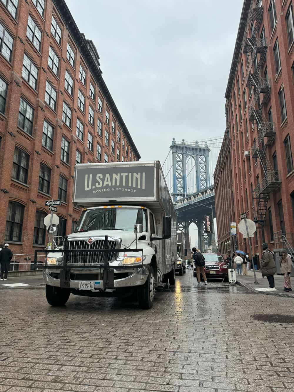 A U. Santini Moving & Storage truck driving on wet cobblestone streets in the DUMBO neighborhood of Brooklyn, with the iconic Manhattan Bridge perfectly framed in the background between red brick warehouse buildings