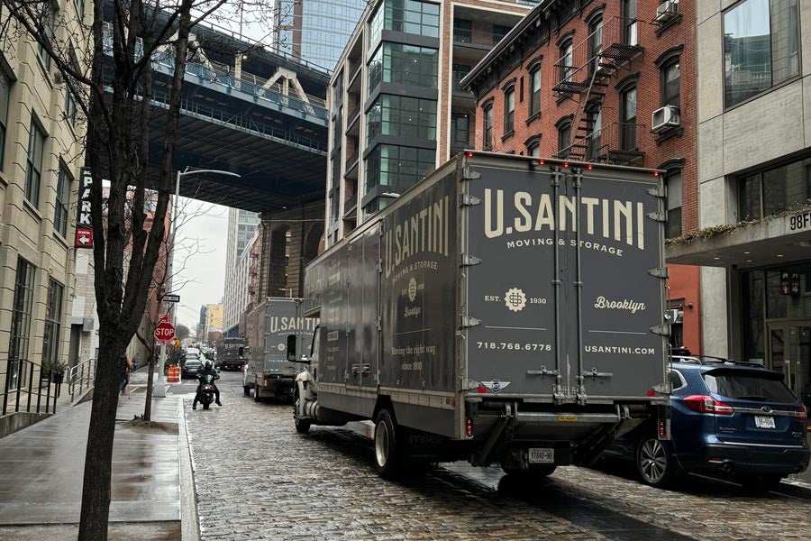 Two U. Santini Moving & Storage trucks parked on a cobblestone street in DUMBO, Brooklyn, with a bridge overhead and the company's website usantini.com and phone number visible on the rear