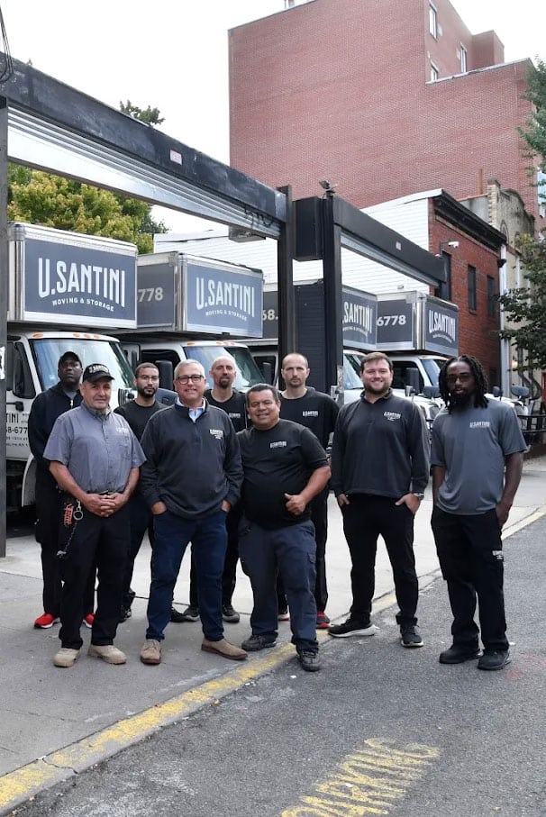 The U. Santini Moving & Storage team of nine movers posing in front of a row of branded white moving trucks on a Brooklyn street